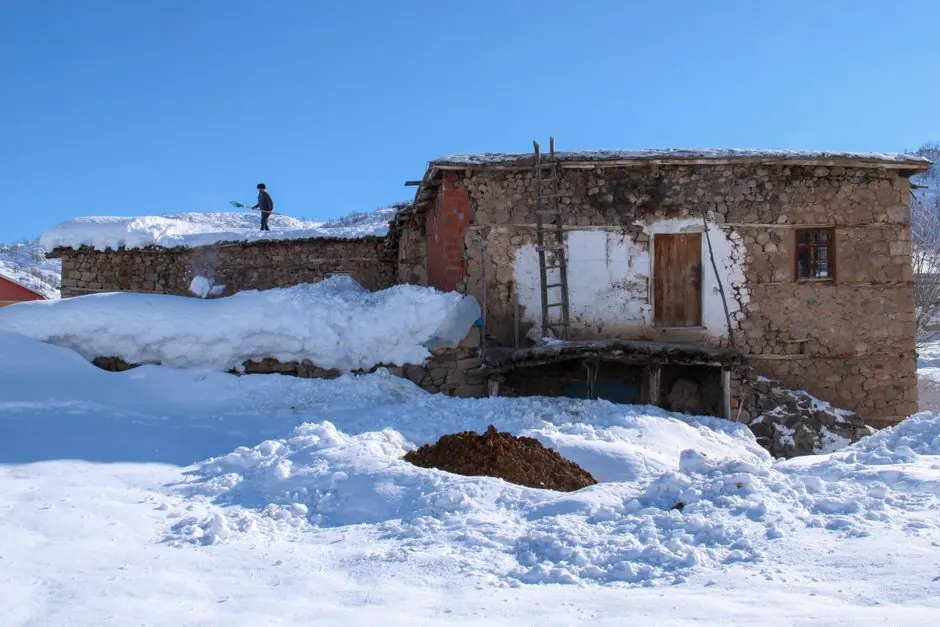 A person shoveling snow on a rustic stone house in a winter landscape.